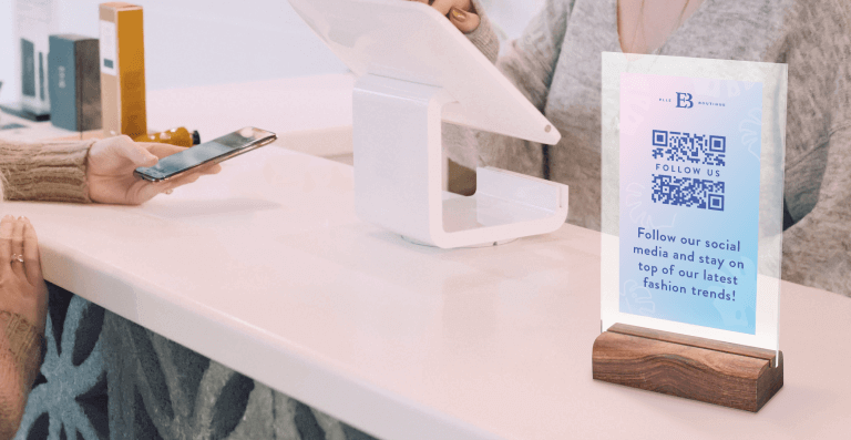 A woman with her hands on the counter next to an acrylic QR Code sign on display to follow Elle Boutique’s social media pages.