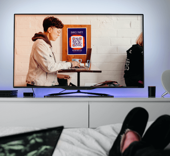 A person wearing slippers watching the television set that is displaying a QR Code ad.