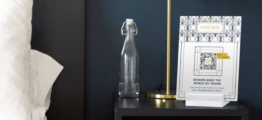 A bedside table holds an empty water bottle, a lamp, and a table display with a Google business review QR Code for a hotel.