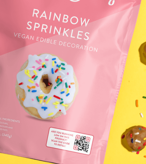 A pink and yellow packaging of sprinkles with a picture of a white glazed donut covered in sprinkles and a QR Code.
